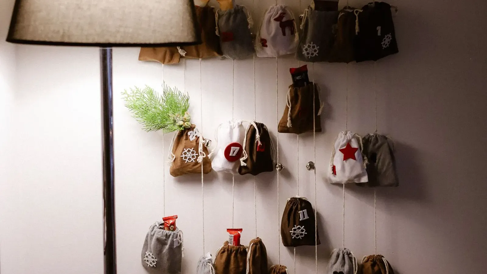 Homemade advent calendar of small fabric pouches hanging from a triangular rail on a wall beside a lit floor lamp