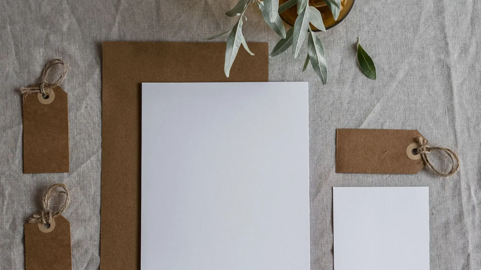 Blank kraft tags, a white card and a brown envelope laid on grey linen with an olive sprig