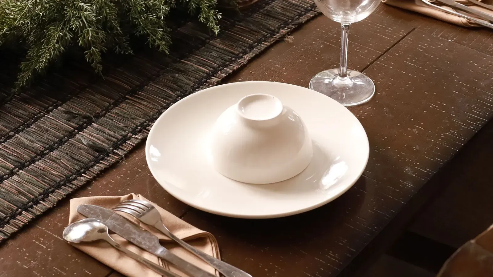Wooden dining table with a fir greenery runner, tall candles and white plates laid for Christmas dinner