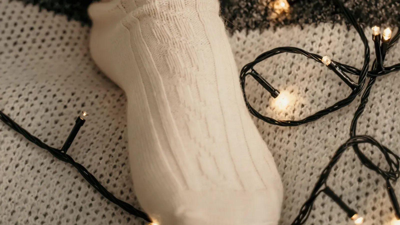 Foot in a cream cable-knit sock resting on a woolly rug among tangled black-wire fairy lights