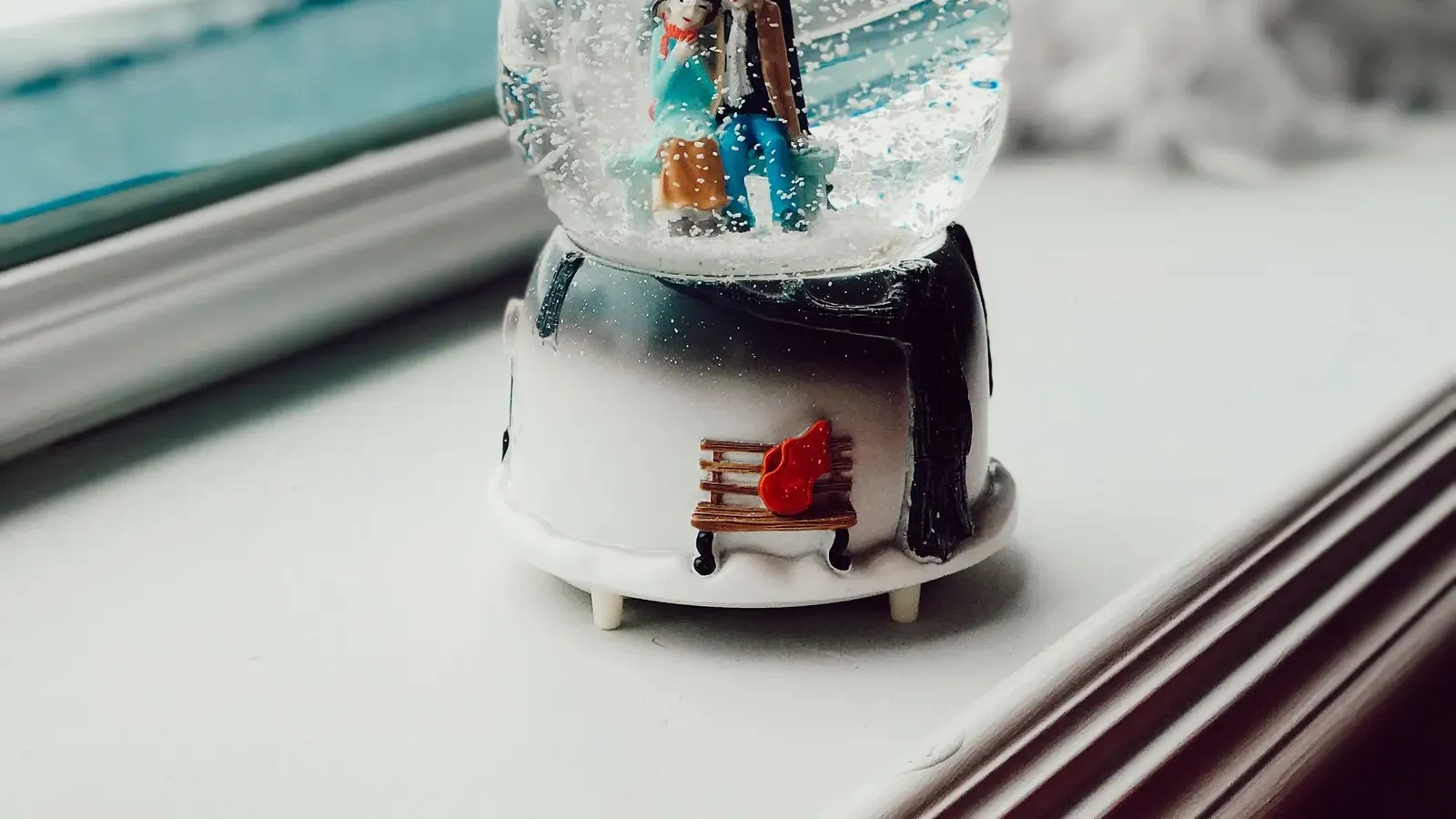 Snow globe with a couple under a lamp post on a white window sill, snowy garden blurred behind the glass