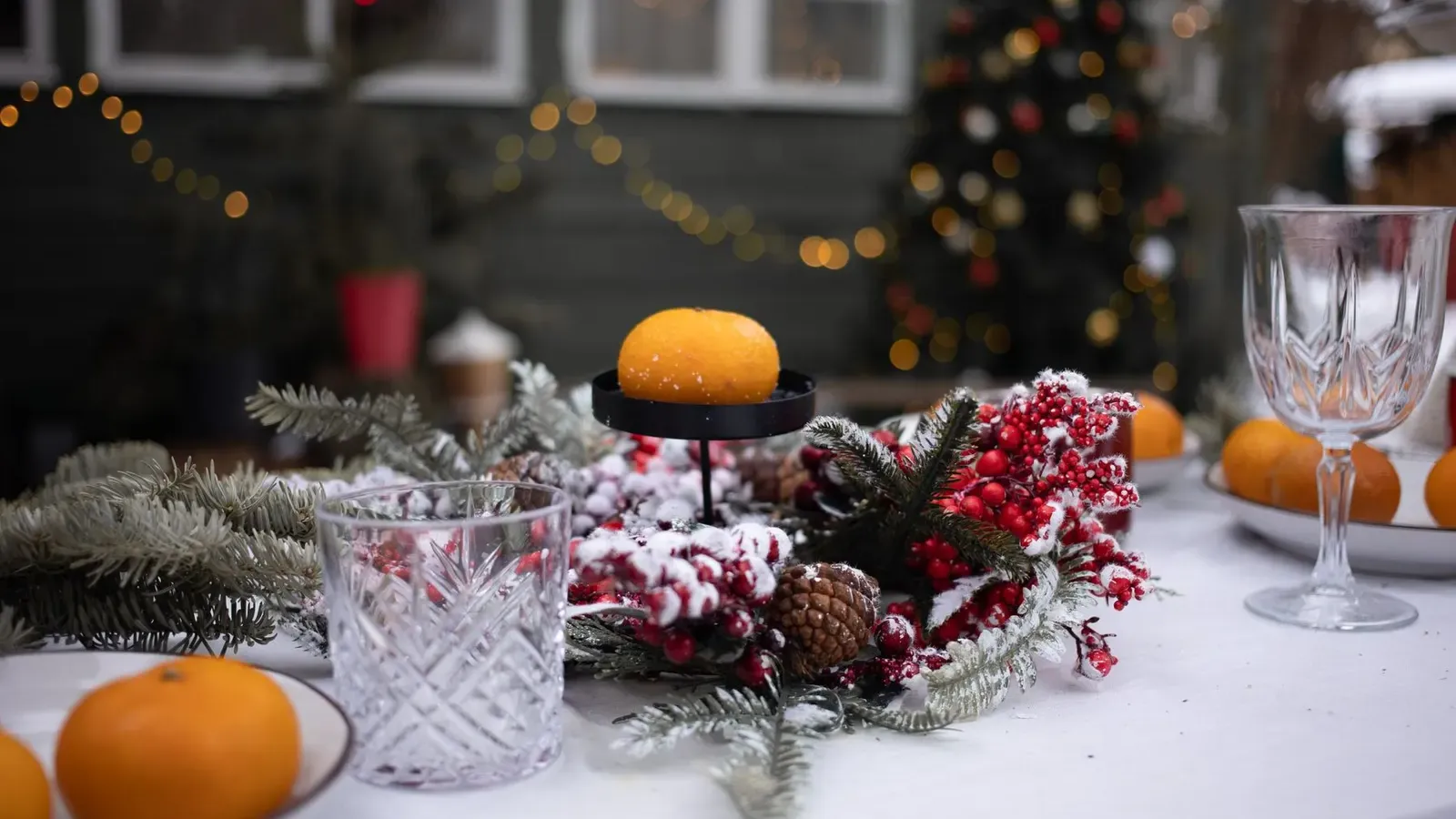 Outdoor table with a snowy berry and pine wreath, tangerines and crystal glasses, a lit tree glowing behind