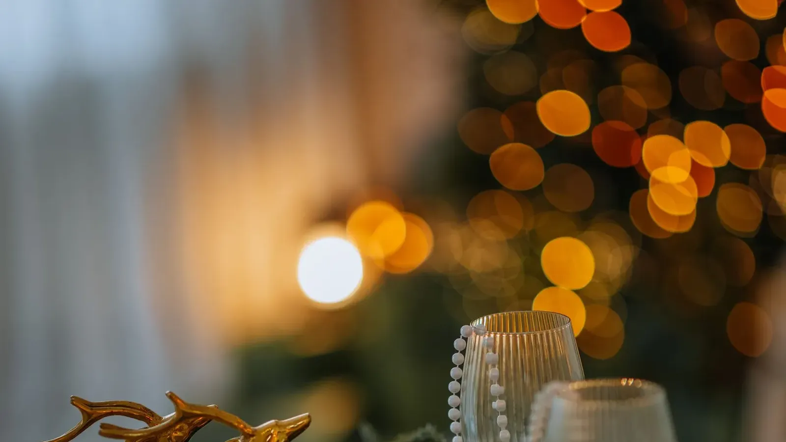 Brass reindeer figures, pearl strands and fir sprigs on a dinner table with warm tree lights blurred behind