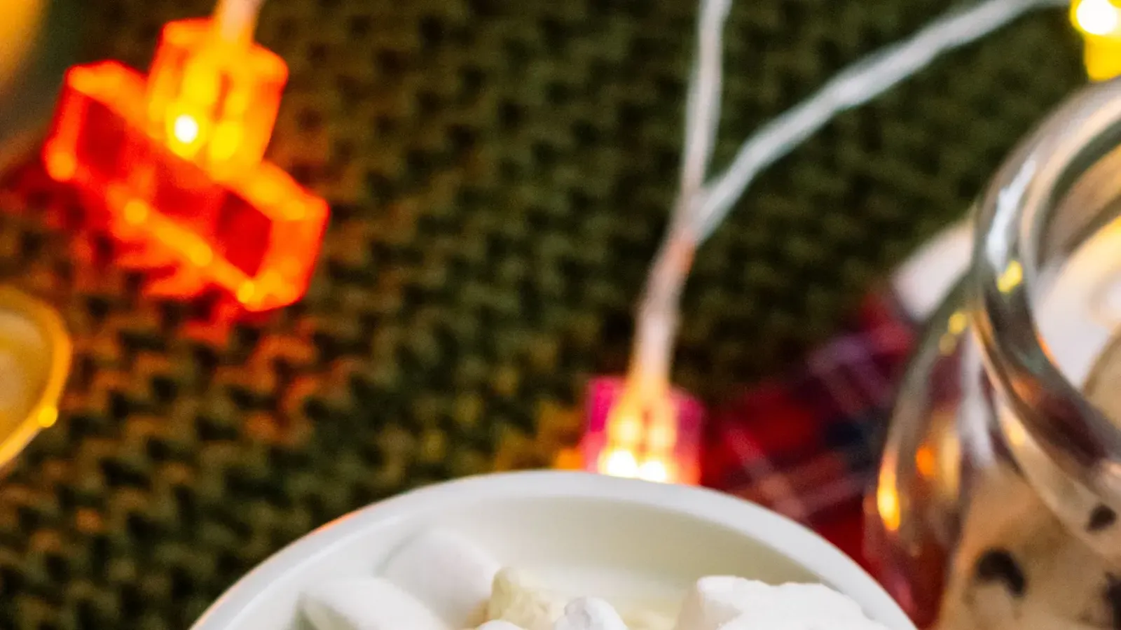 Snowman mug of hot chocolate topped with snowflake-shaped marshmallows, with fairy lights and a knitted cloth behind