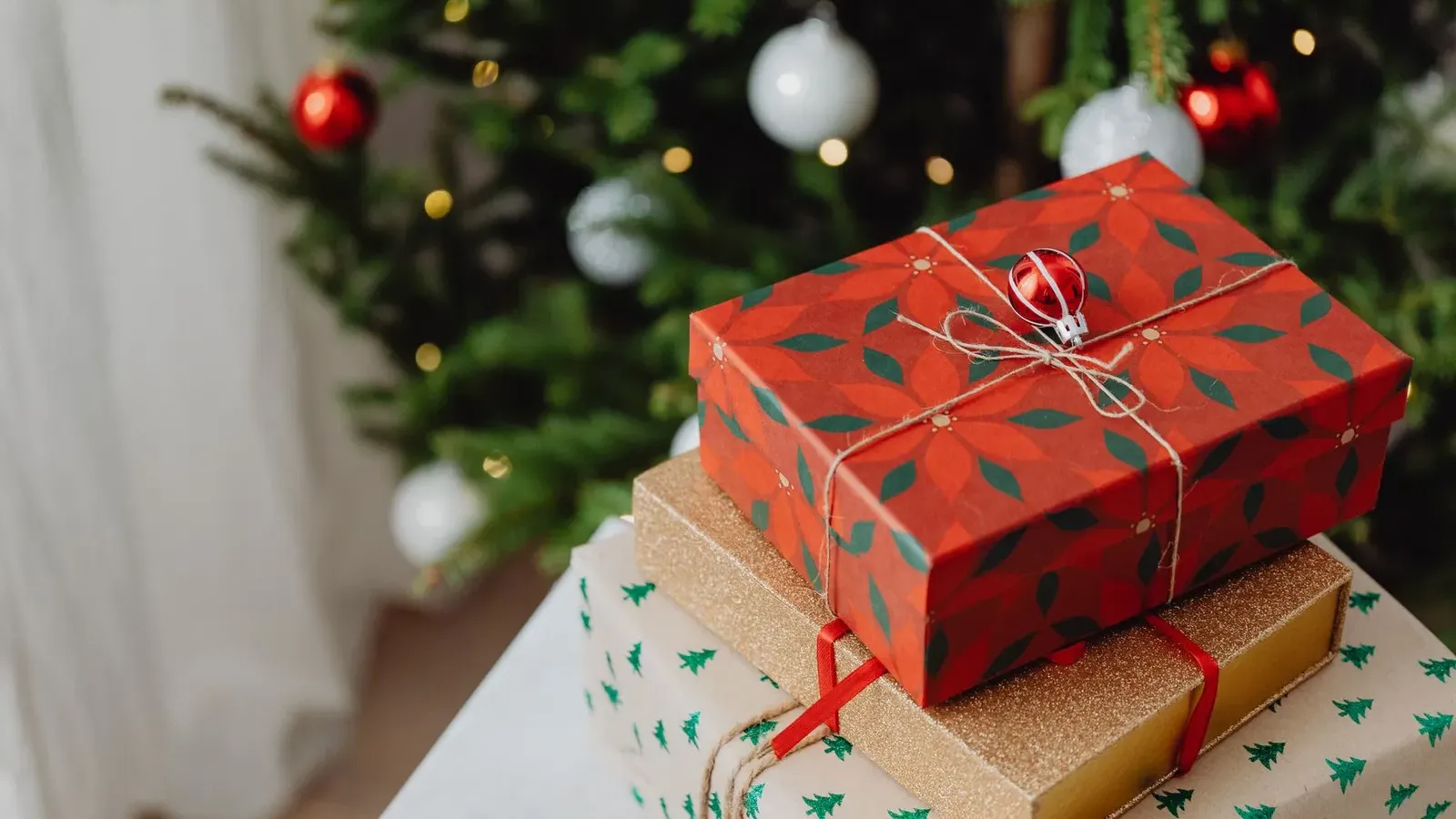 Red parcel with leaf-pattern paper and a bauble on the string, stacked on gold and tree-print gifts before a tree