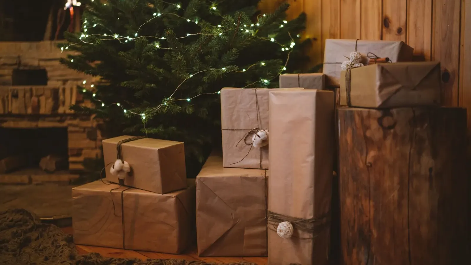 Kraft paper parcels tied with twine stacked beside a fairy-lit fir tree in a room with wooden walls
