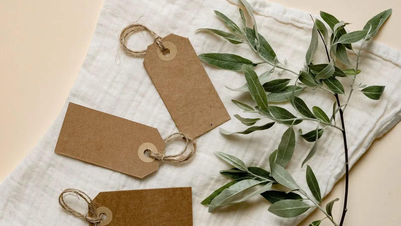 Three blank kraft gift tags with twine loops on white muslin beside an olive branch