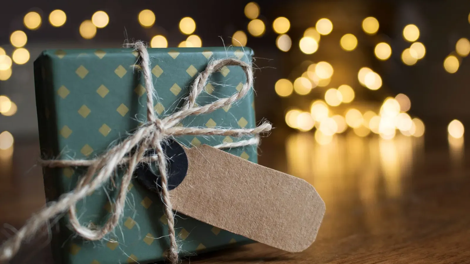 Small green present with a gold diamond pattern, natural twine and a blank kraft tag, warm bokeh lights behind