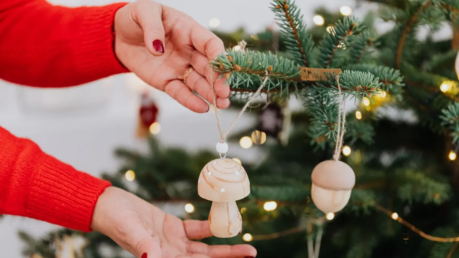 Woman's hands in a red jumper hanging wooden mushroom and acorn ornaments on a fir branch with fairy lights