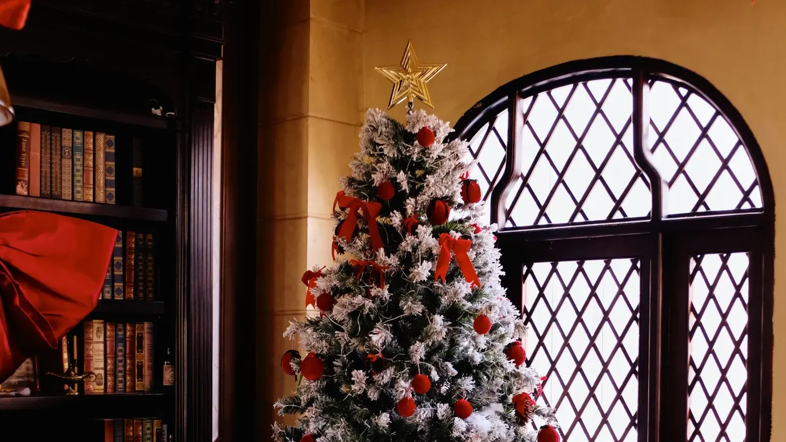 Frosted Christmas tree with red bows and baubles beside a leaded arched window in a book-lined room