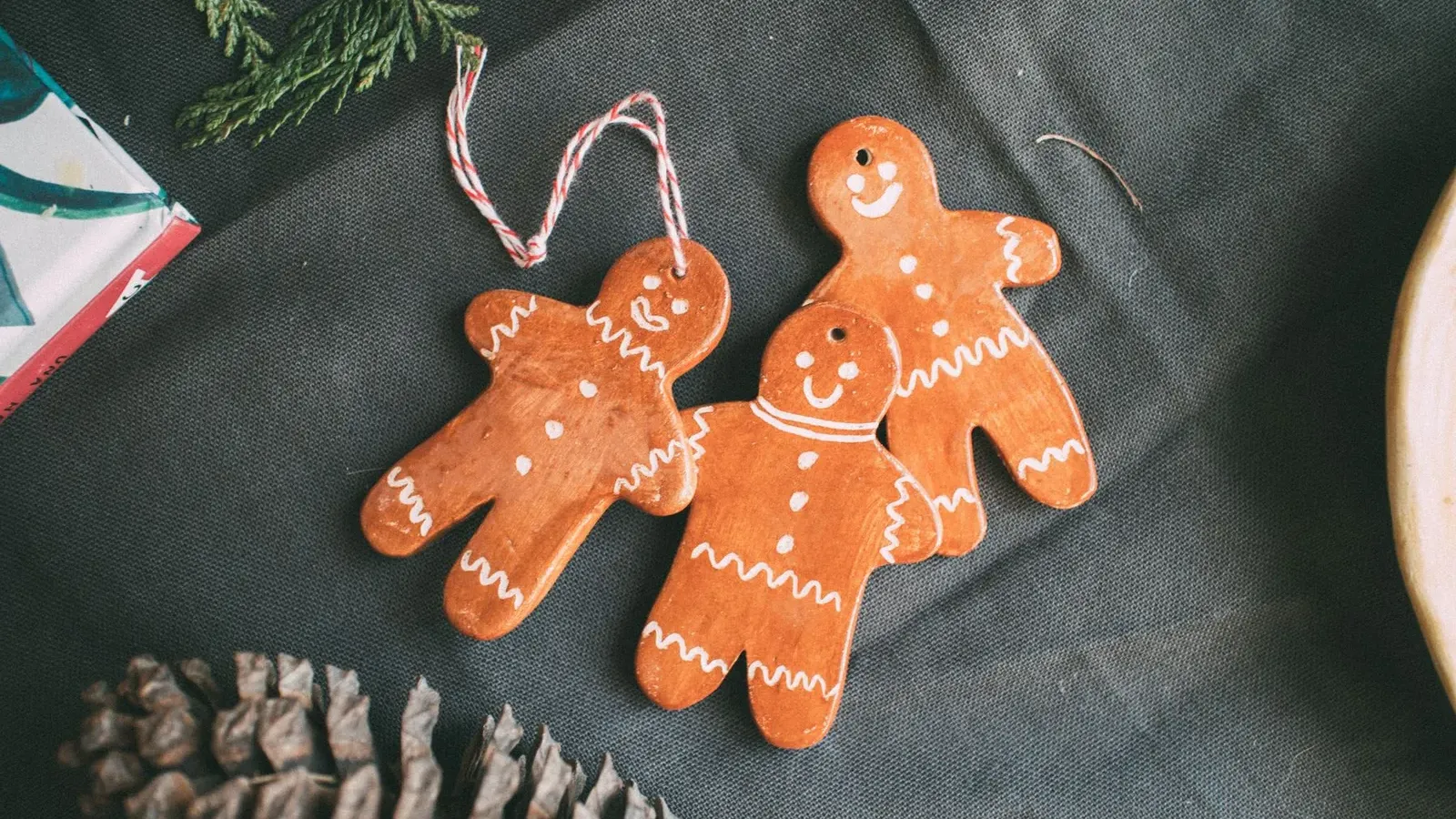 Three salt-dough gingerbread men ornaments with white icing lines on a dark cloth beside a pine cone and fir sprig