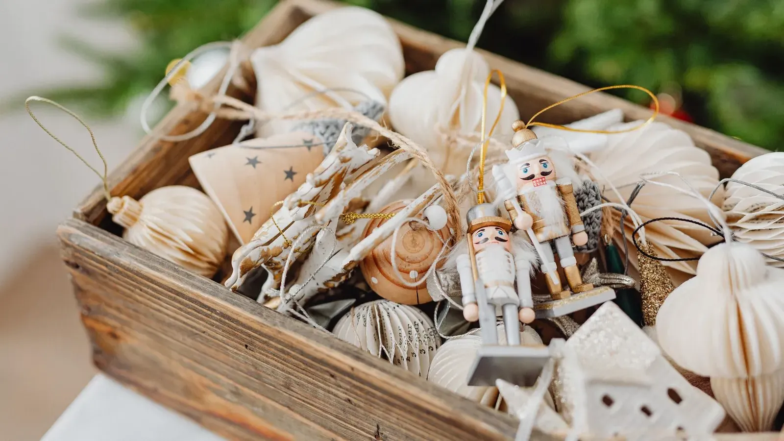 Wooden crate filled with white paper honeycomb ornaments, small white nutcracker figures and tiny houses, tree lights blurred behind