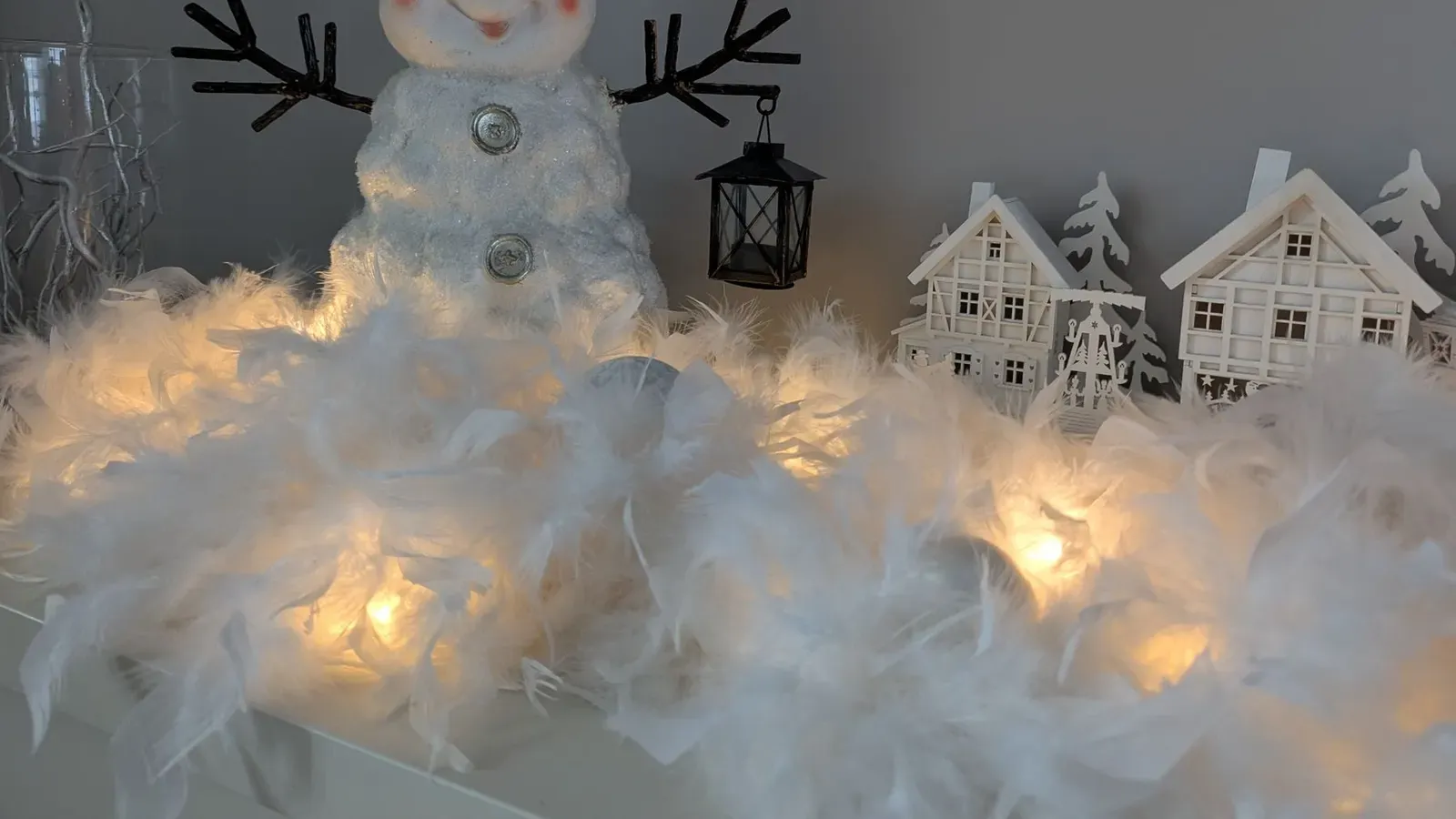 Smiling snowman figure with a small lantern sitting on glowing white feathers, with little wooden houses beside