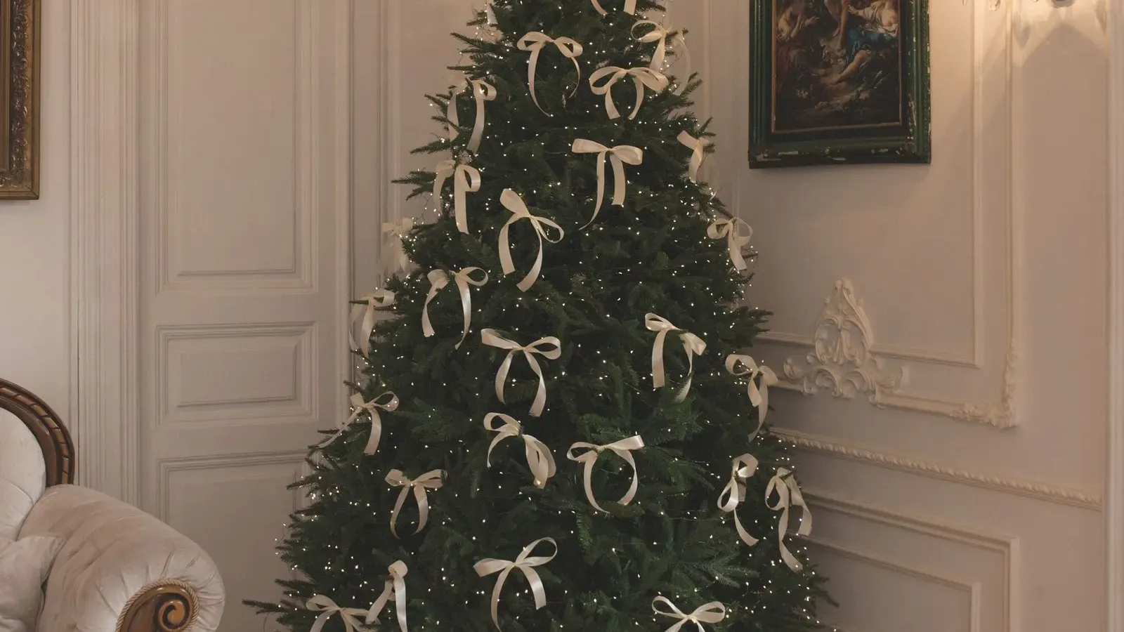Christmas tree with cream ribbon bows in a corner of a panelled room with parquet floor, wall sconce and painting