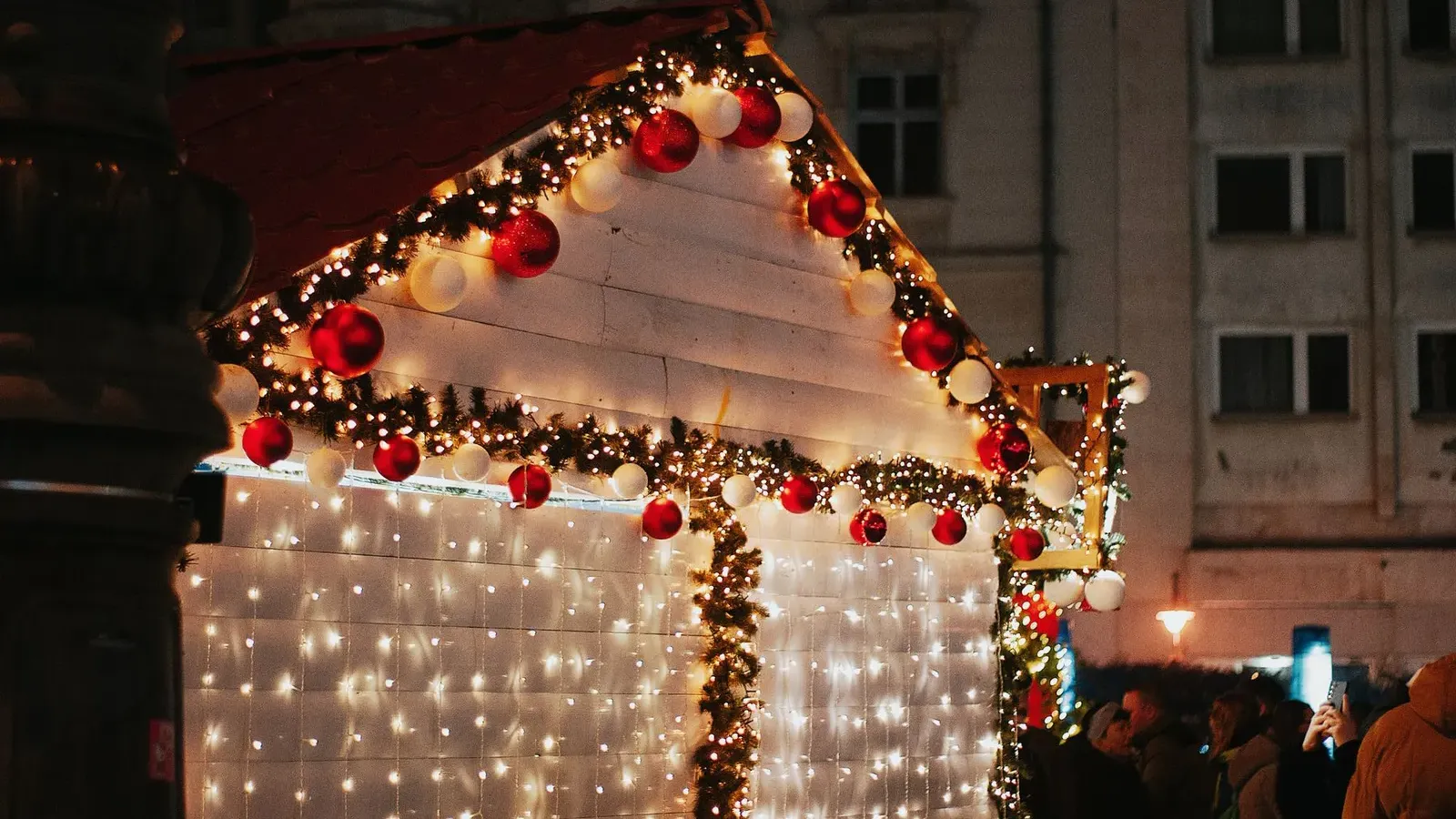 White market hut hung with curtain lights, a bauble garland and fir boughs, with a small crowd at night