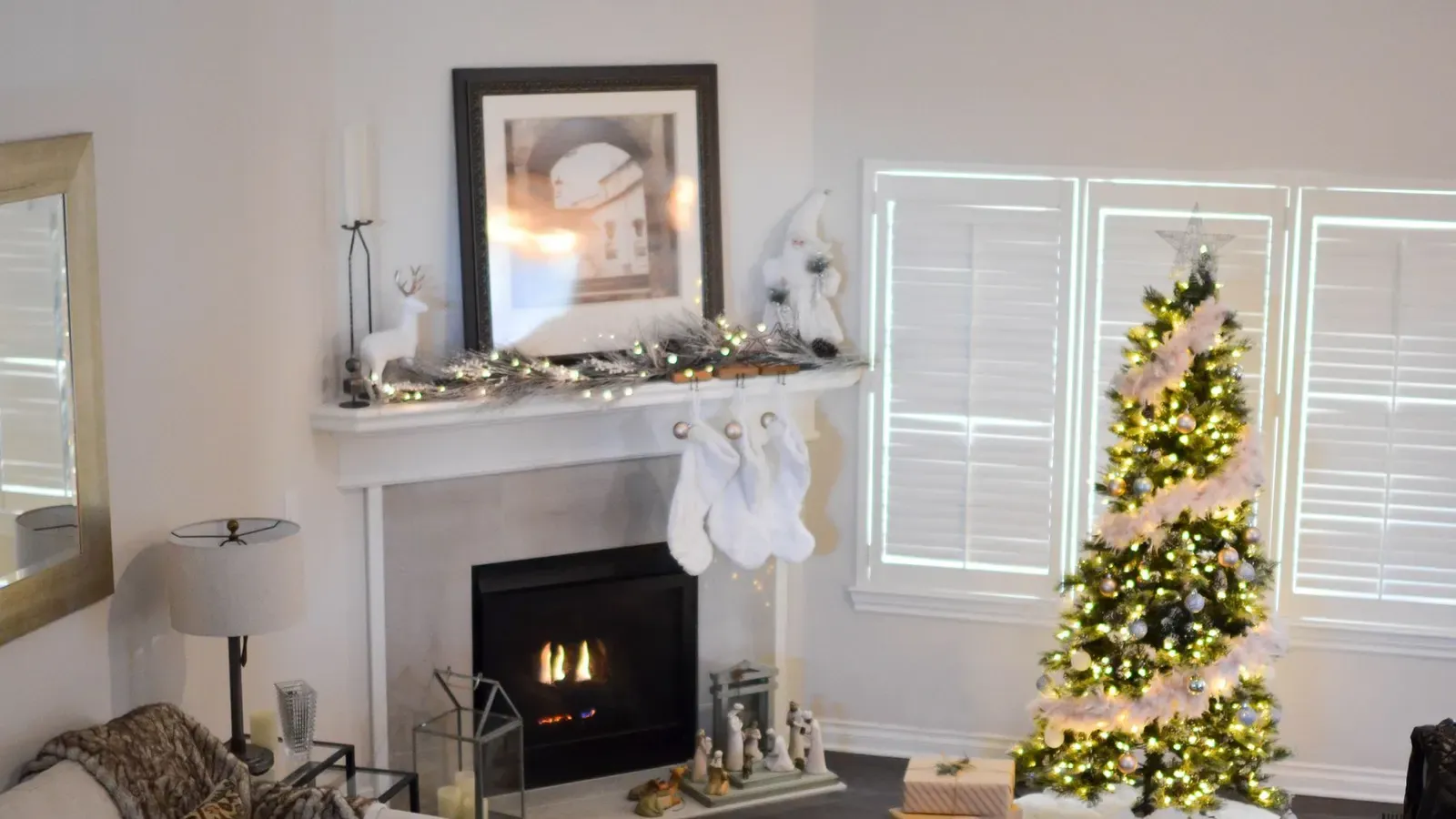 Bright living room with a lit tree draped in white tinsel, white stockings on a white fireplace and shuttered windows