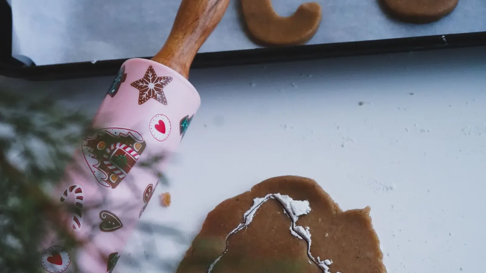 Rolled gingerbread dough with a tree-shaped cutter and a pink patterned rolling pin, baked cookies on a tray above