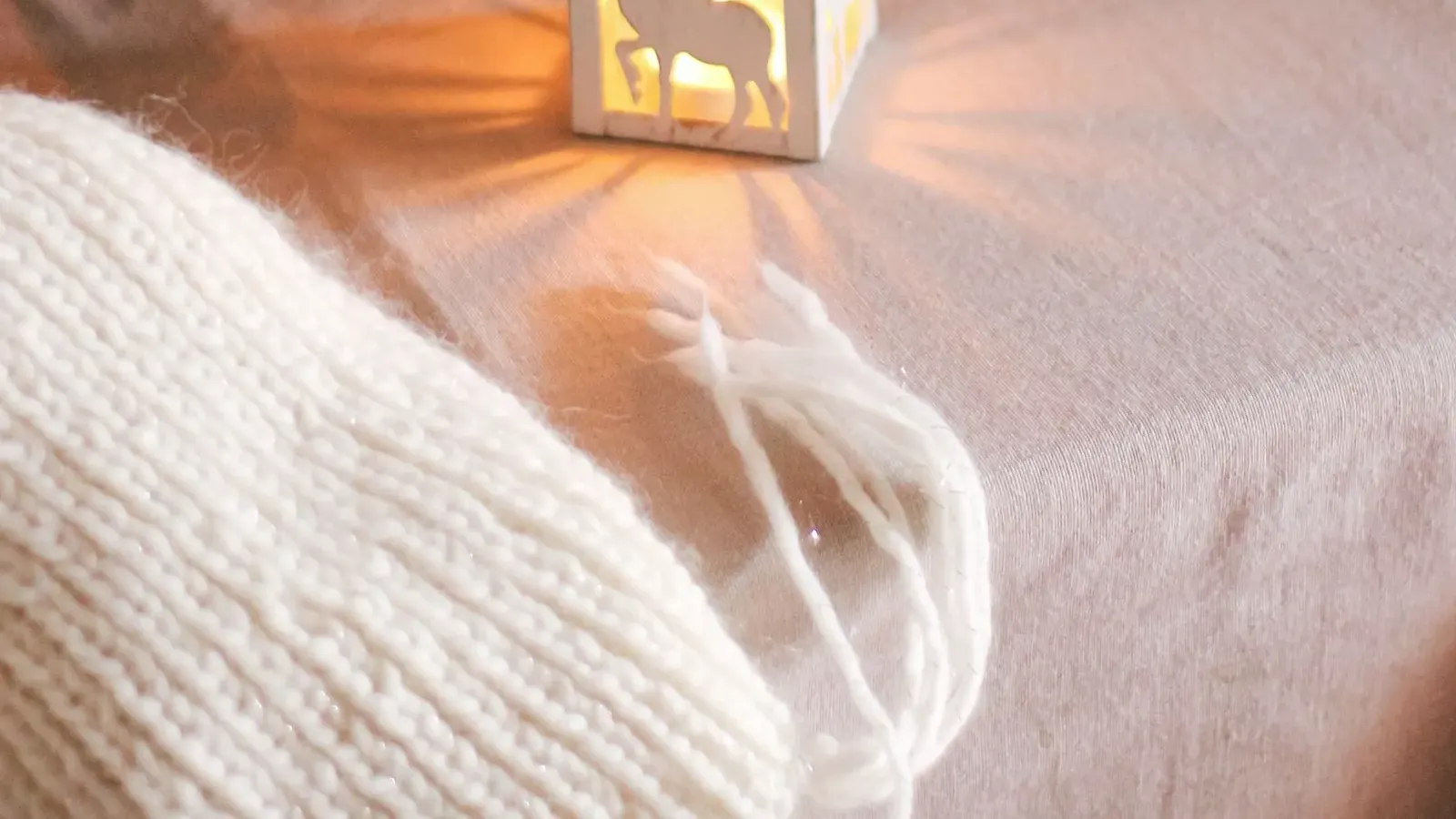 Small white lantern with a reindeer cut-out glowing on a linen cloth beside a chunky knit throw and greenery