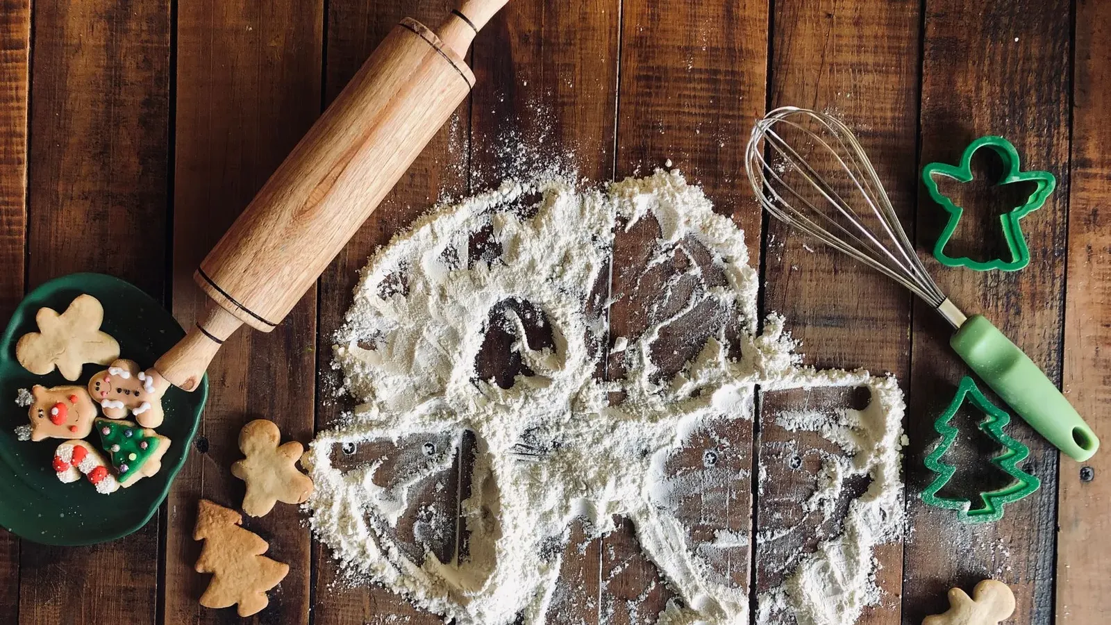 Flour angel drawn on a dark wooden table with a rolling pin, whisk, green cutters and small gingerbread biscuits