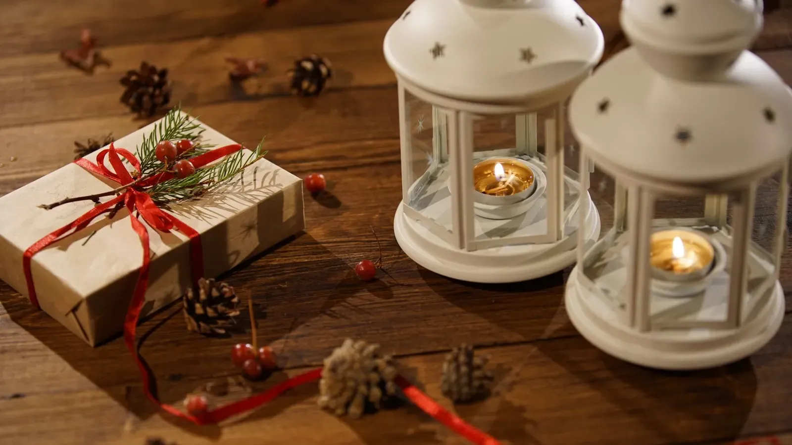 Two white lanterns with lit tealights beside a kraft gift with red ribbon and pine cones on a wooden table