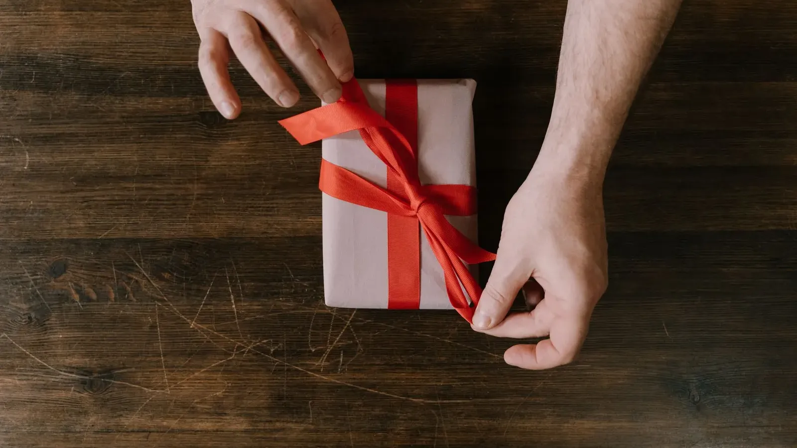 Two hands tying a red ribbon around a small white gift box on a dark wooden table, seen from above