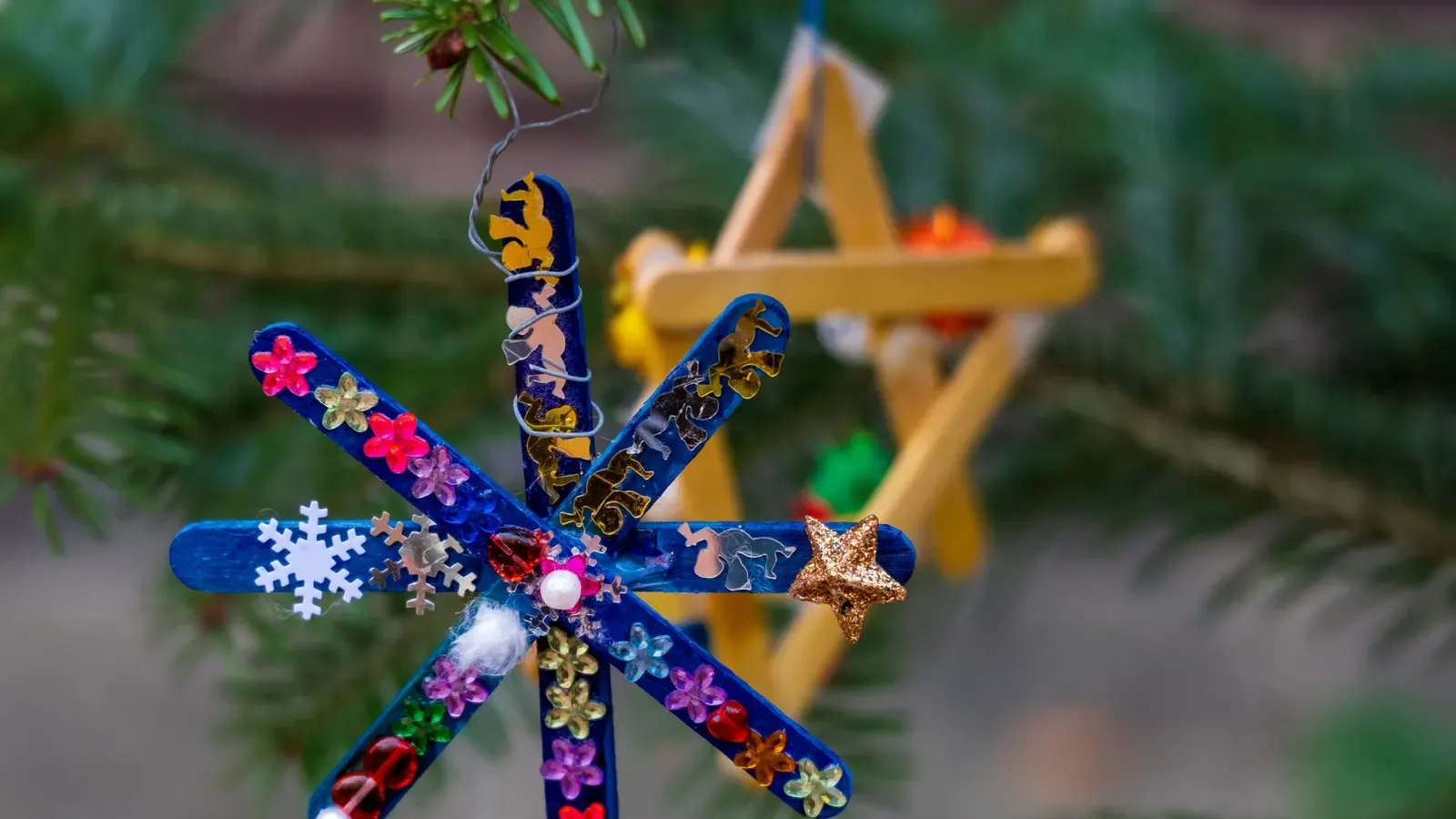 Child-made snowflake ornament of blue lolly sticks with sequins and a wooden star on a fir branch