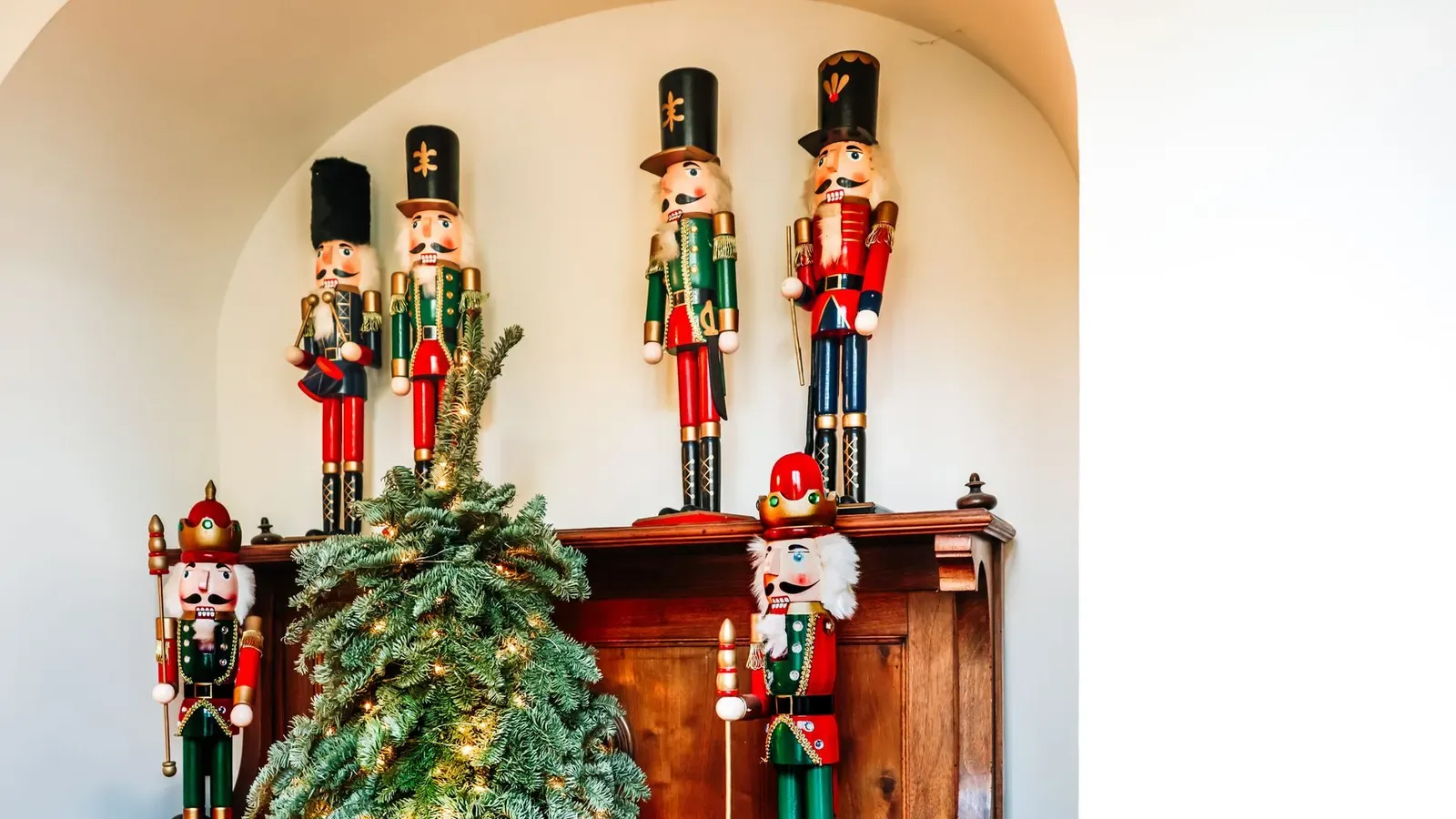 Six painted wooden nutcrackers on a wooden cabinet beside a small lit fir tree in a white alcove