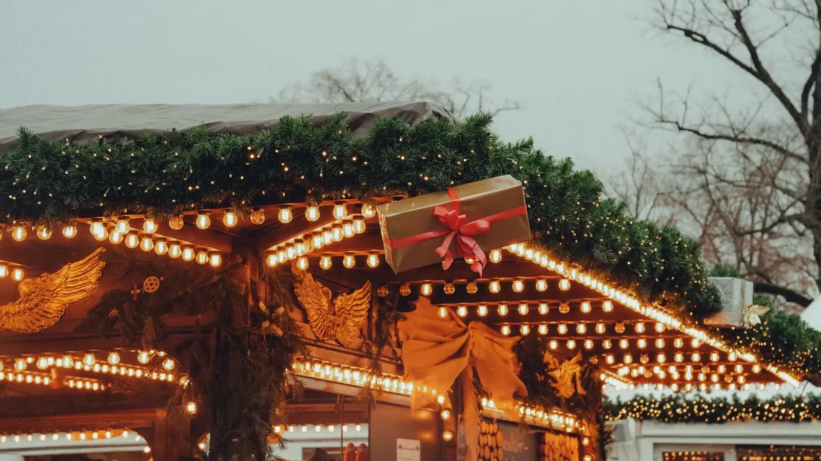 Wooden market stall roof outlined with warm bulbs and fir garland, topped with a large wrapped gift