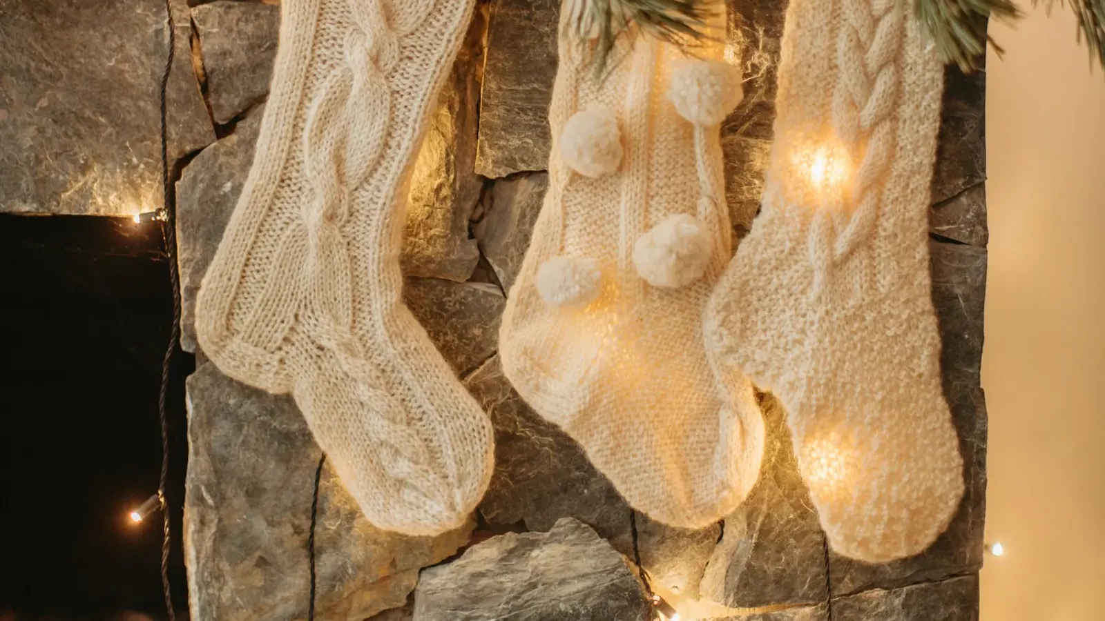 Three cream knitted stockings hanging on a stone fireplace under a fir branch with warm fairy lights
