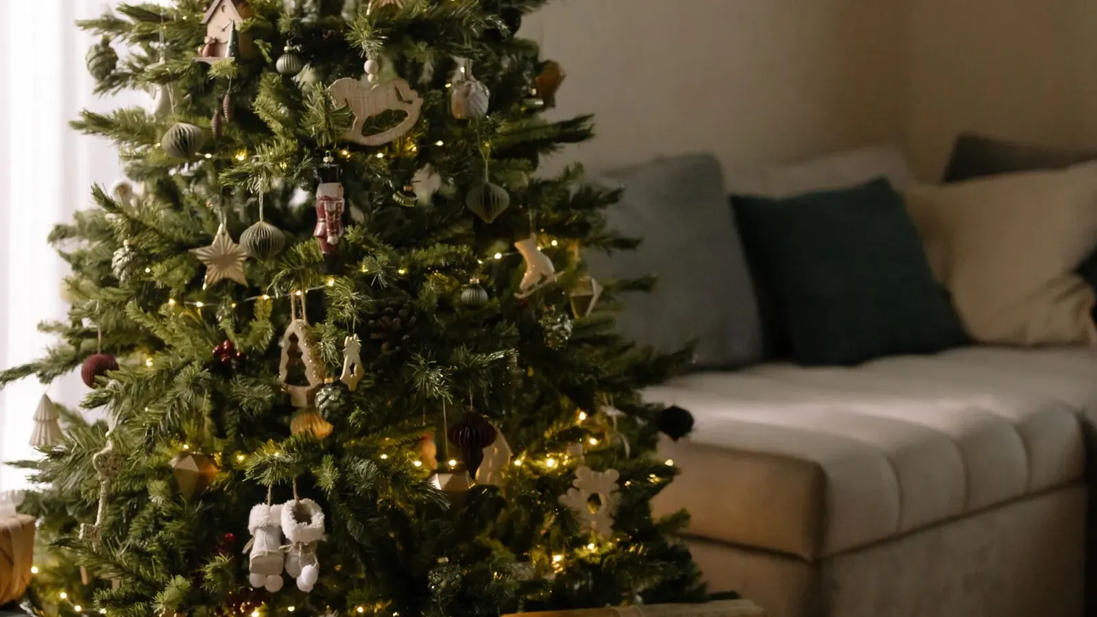Christmas tree with white ornaments and small lights beside a sofa, gifts on a mustard rug, wreath on the wall