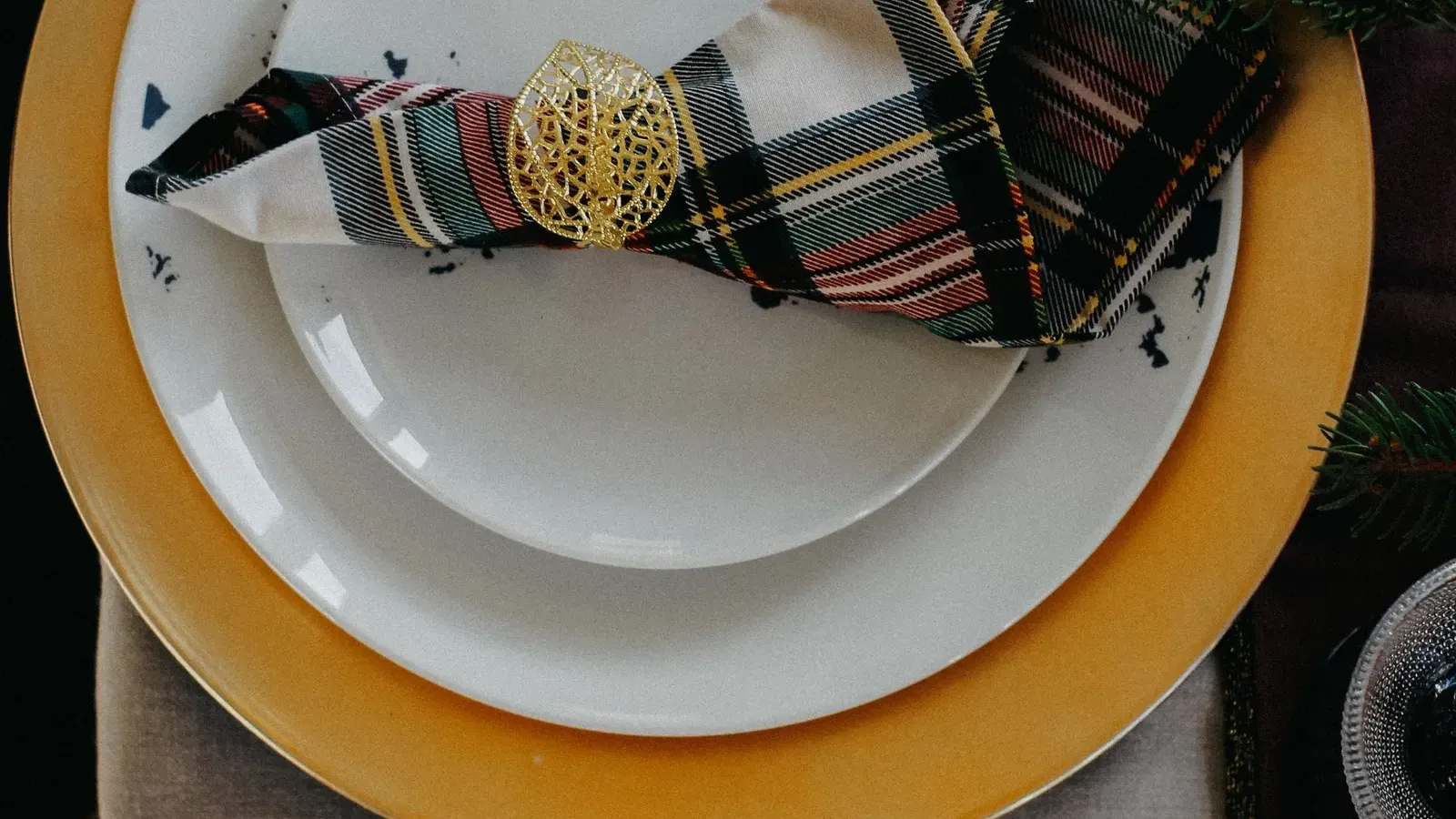 Tartan napkin in a gold ring with a fir sprig on white plates stacked on a gold charger, gold cutlery